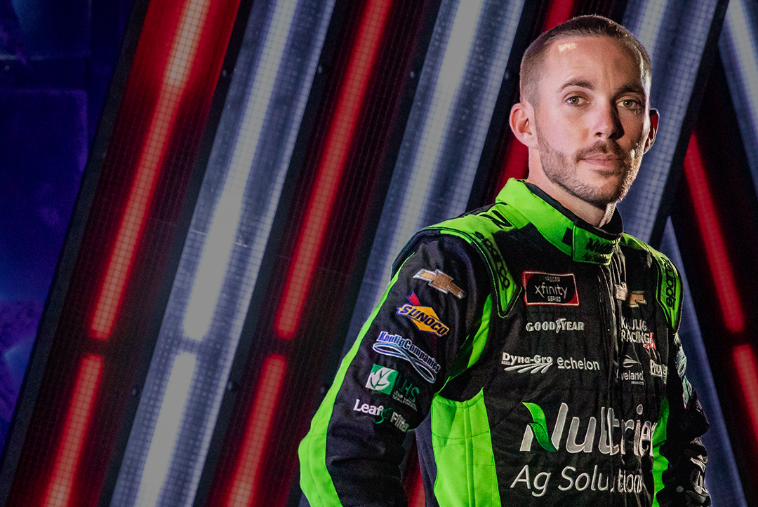 NASCAR driver Ross Chastain poses with his hands on his hips in front of a blue and red background. He is wearing a black and green Nutrien Ag Solutions racing jacket.