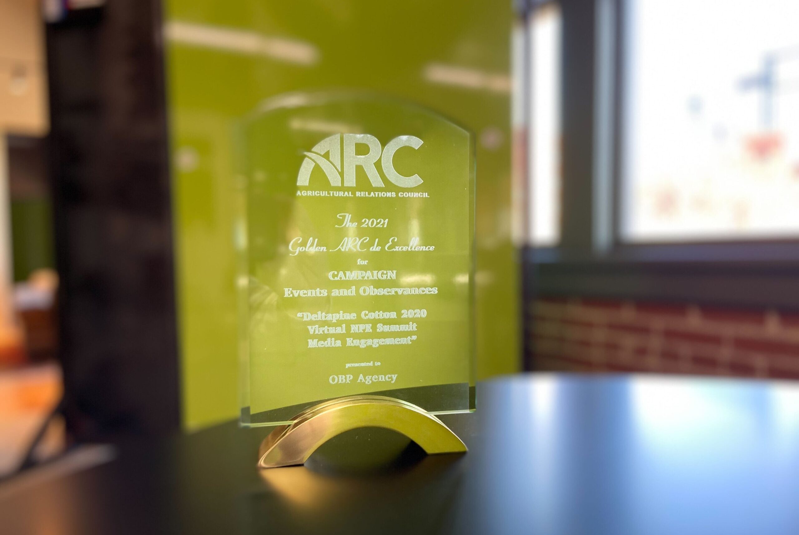 An Agricultural Relations Council ad sits on a table in an office building. The award reads: The 2021 Golden ARC de Excellence for Campaign Events and Observances. "Deltapine Cotton 2020 Virtual NPK Summit Media Engagement" presented to OBP Agency.