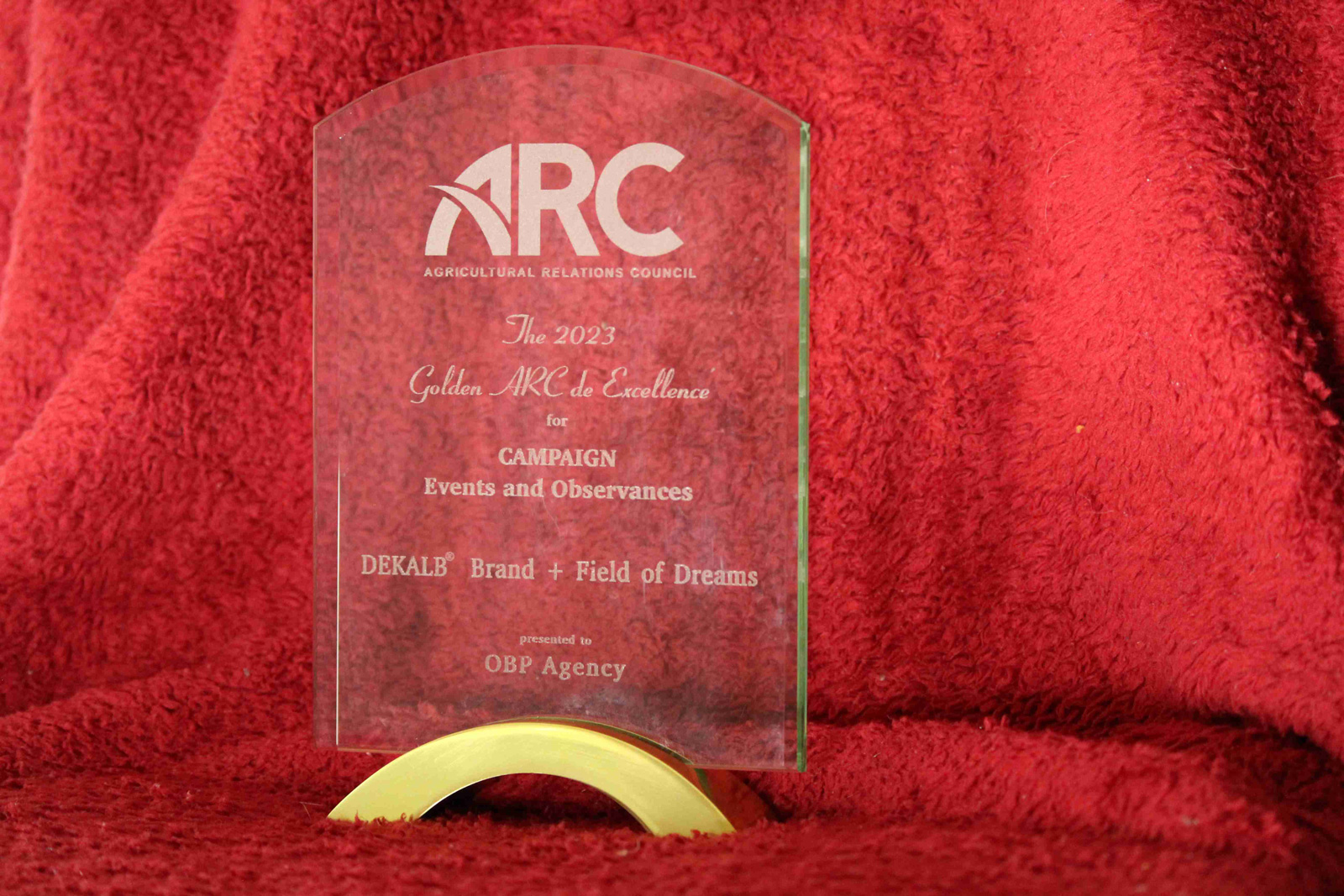A 2023 Golden ARC de Excellence award sits on a red blanket. The award is for Campaign Events & Observances for DEKALB Brand + Field of Dreams presented to OBP Agency.